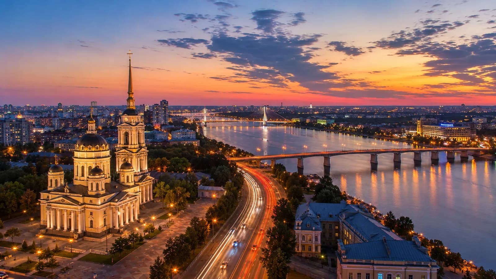 Благовещенский собор с золочёным шпилем на возвышенном правом берегу Воронежского водохранилища на закате: мосты через воду, огни города и следы фар на набережной дороге