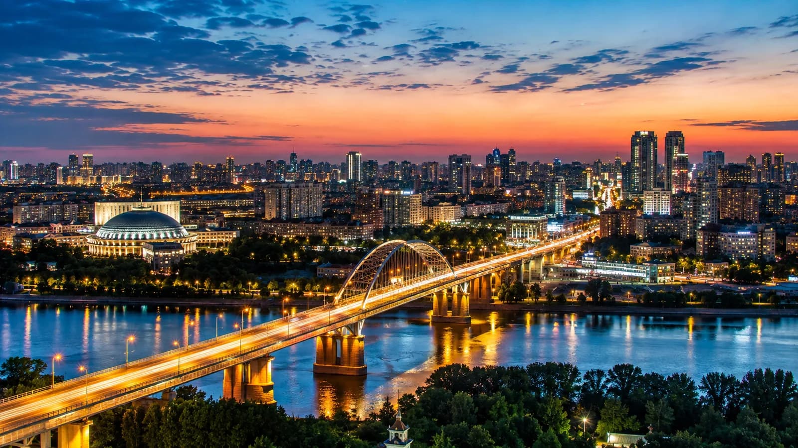 Подсвеченный мост через Обь в Новосибирске на закате, купол театра оперы и балета на набережной и плотная высотная застройка на горизонте