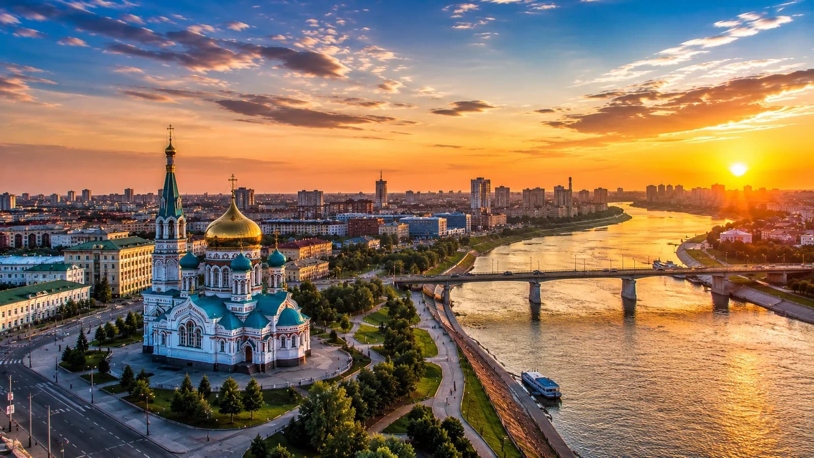 Успенский кафедральный собор с золотым куполом и бирюзовыми шатровыми крышами на берегу Иртыша в Омске на закате — вид с высоты на изгиб реки, мост и городскую застройку до горизонта